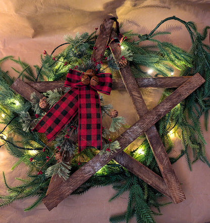 Decorative wreath with greenery, wooden planks, and a red and black checkered bow on a neutral background.