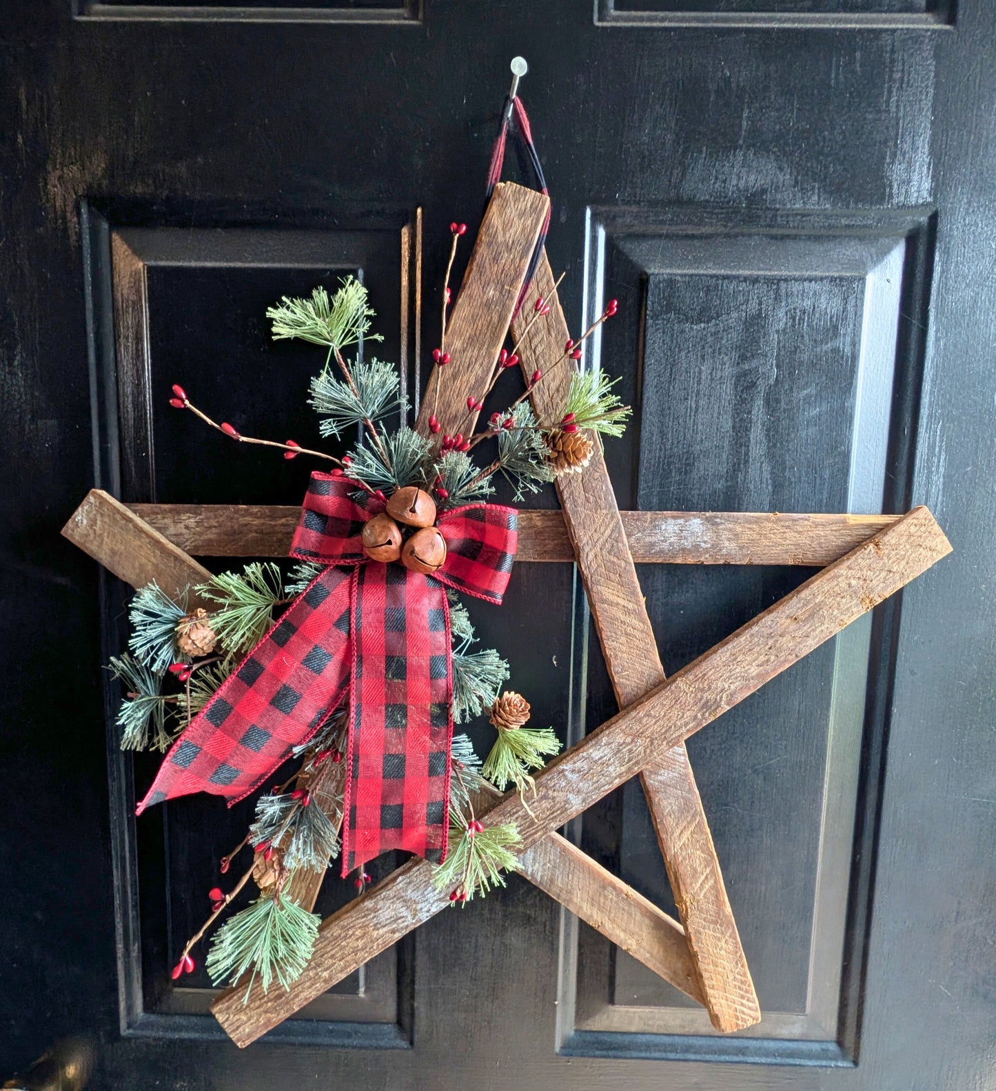 Wooden star decoration with greenery and a red plaid bow on a black door.