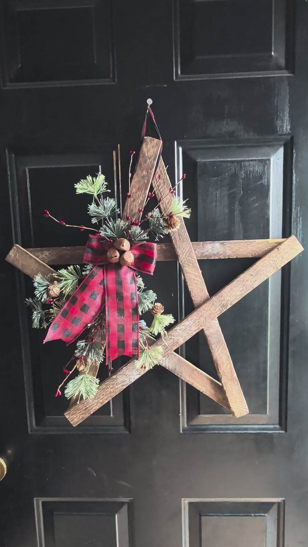 Video of a rustic wooden star wreath hanging by a buffalo plaid holder, has greenery 3 bells, and long buffalo plaid print on the front. Hanging on a black door.
